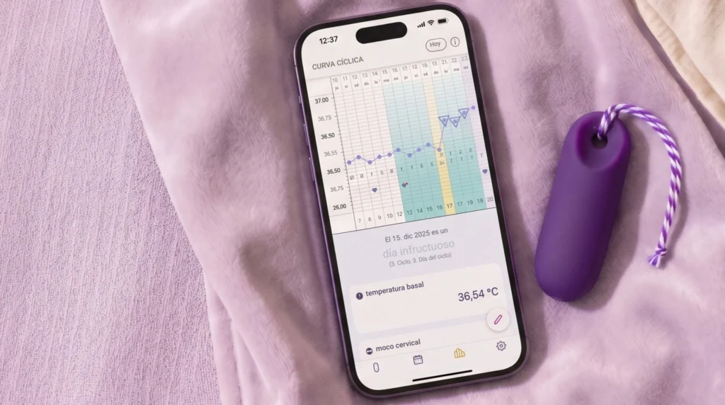 Hay un smartphone junto al pequeño sensor Trackle de color lila sobre una manta de color violeta claro. En la pantalla se ve una aplicación para el ciclo menstrual con una gráfica de la temperatura basal y el texto «día infértil».