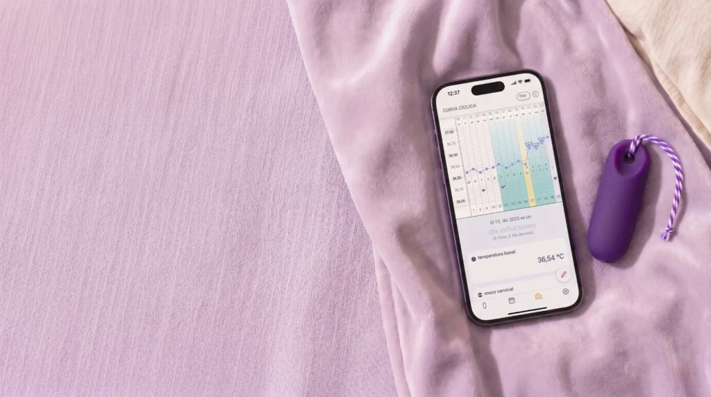 Hay un smartphone junto al pequeño sensor Trackle de color lila sobre una manta de color violeta claro. En la pantalla se ve una aplicación para el ciclo menstrual con una gráfica de la temperatura basal y el texto «día infértil».