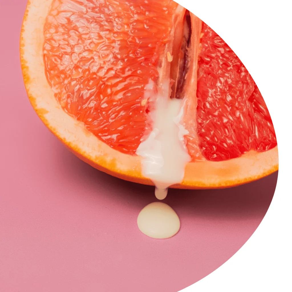 Primer plano conceptual en un corte circular que muestra la mitad de una toronja o pomelo rosa colocada sobre un fondo rosa, con un líquido espeso de color blanco lechoso que gotea desde el centro de la fruta hacia la superficie, utilizado comúnmente como metáfora visual de la anatomía femenina, la salud sexual o el moco cervical (flujo vaginal).