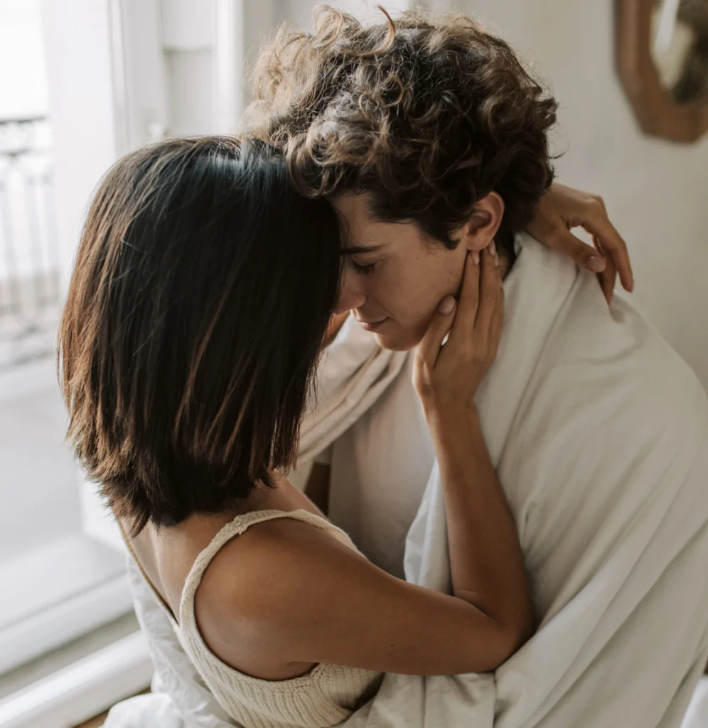 Una pareja joven en un tierno abrazo en el dormitorio. La mujer tiene el pelo corto y oscuro; el hombre, el pelo rizado, y ambos están envueltos en una manta blanca. Se acarician suavemente la cara.