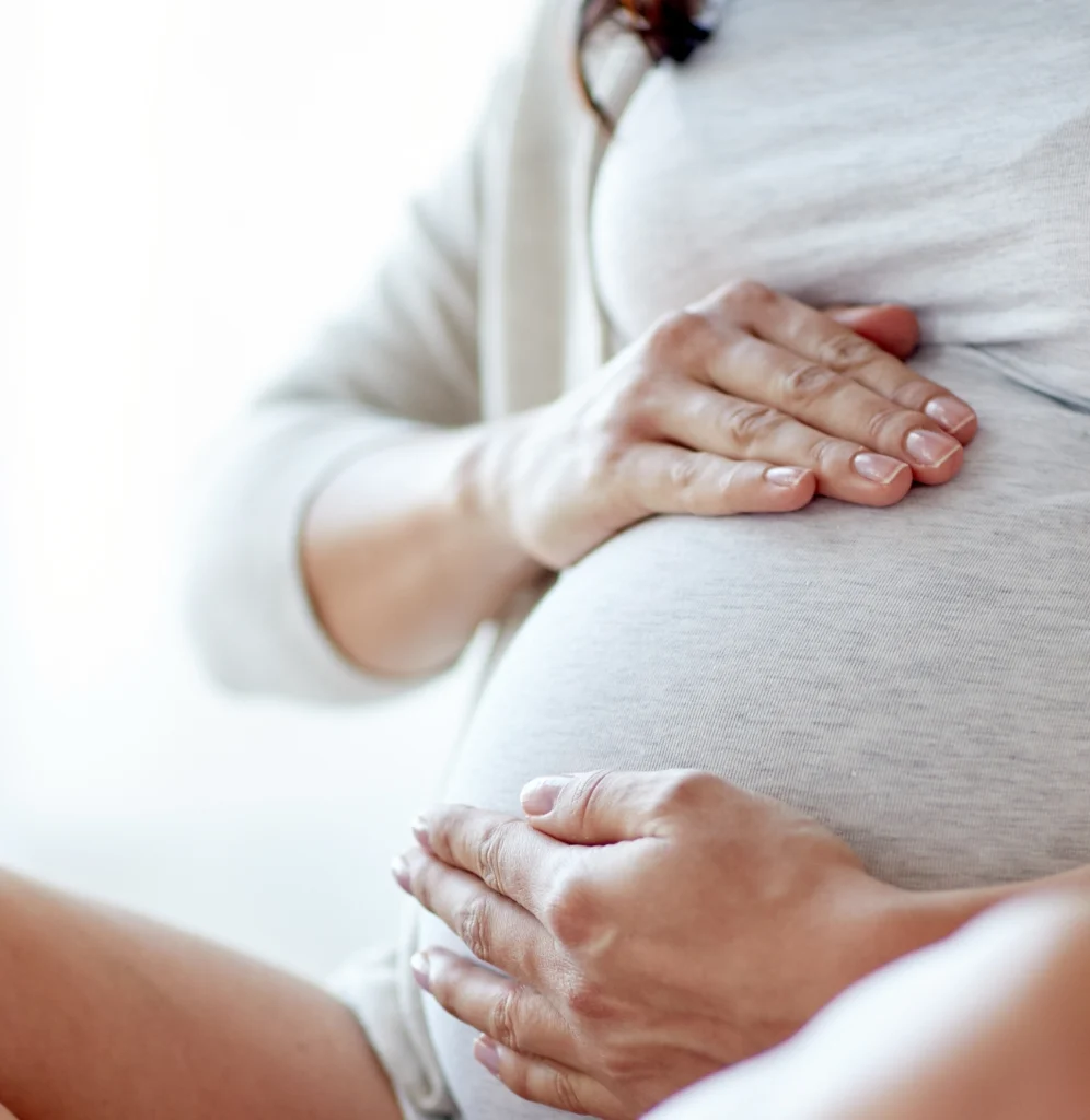 Primer plano de una mujer embarazada vestida con ropa clara, que apoya con delicadeza ambas manos sobre su redondeada barriga.