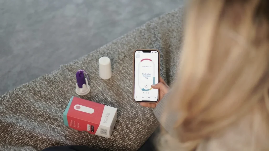 Una vista por encima del hombro de una mujer que sostiene su smartphone. En la pantalla de la aplicación trackle se ve el mensaje «Hoy es un día fértil». Junto a ella, sobre una superficie texturizada, se encuentran el sensor morado de trackle en su soporte y el embalaje del producto. La imagen ilustra el análisis digital del ciclo en la práctica diaria.