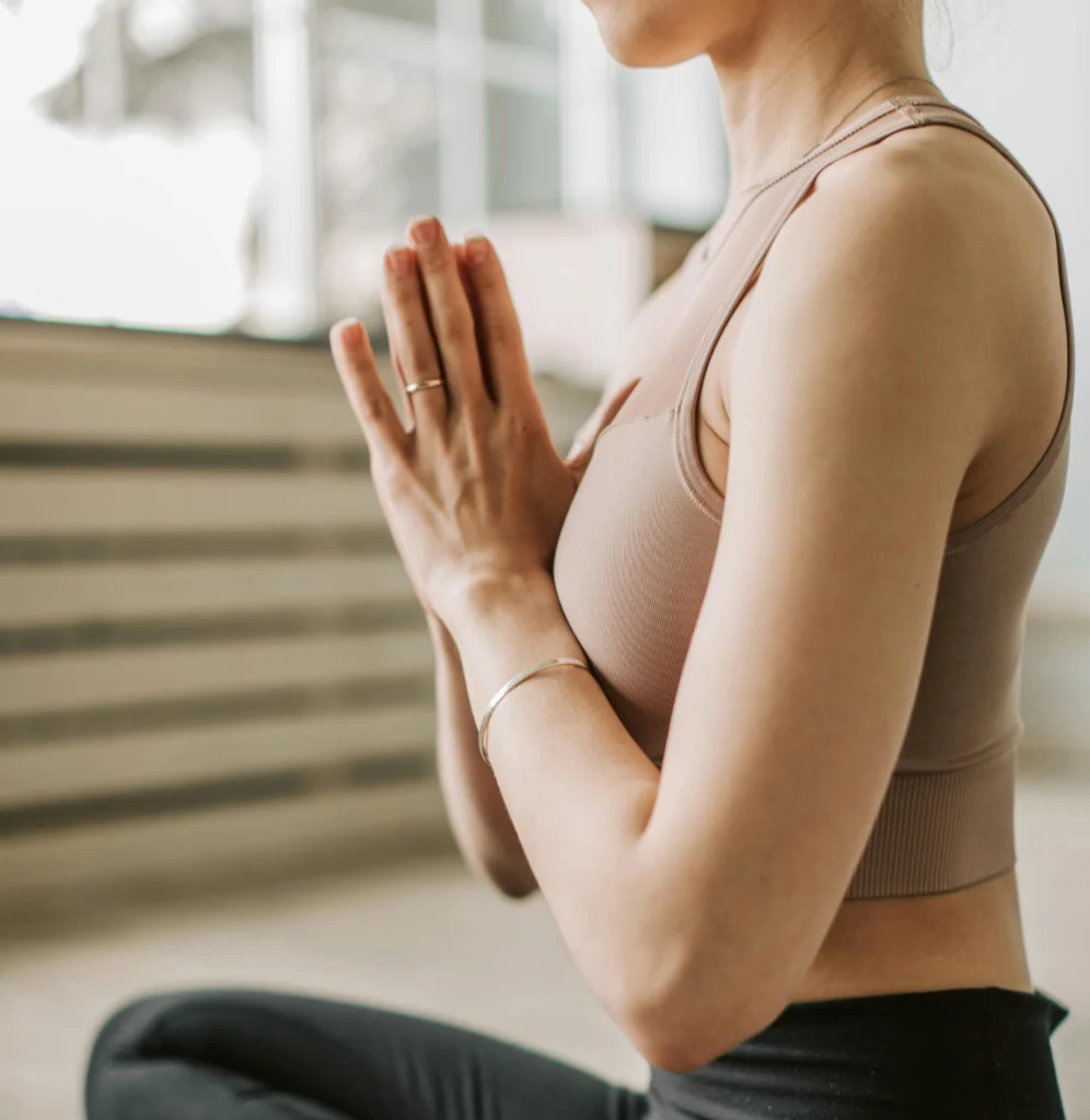 Vista lateral de una mujer en postura de yoga, con las manos juntas delante del pecho en señal de saludo (namasté). Lleva una camiseta deportiva de color beige.