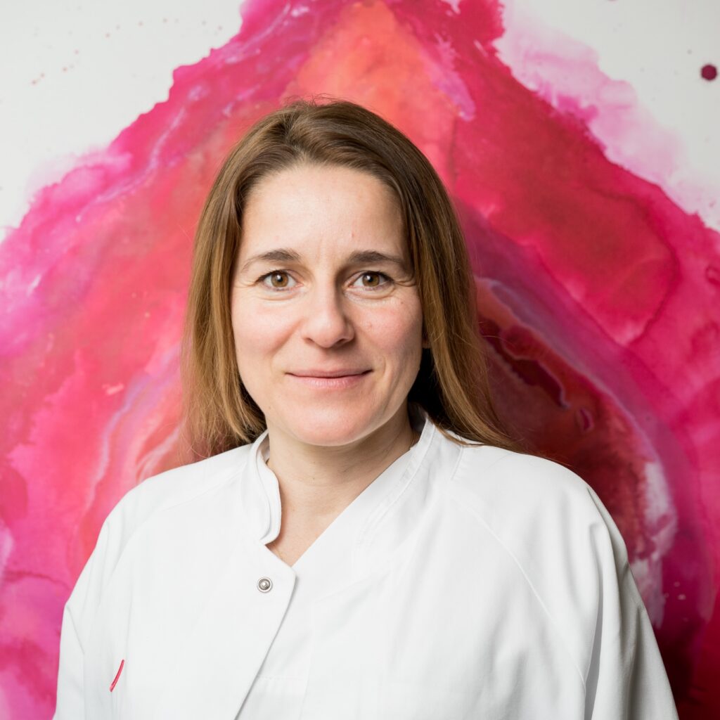 Retrato de la Dra. Mandy Mangler, jefa del servicio de ginecología y obstetricia, con bata blanca de médico ante un fondo pintado en acuarela en tonos rosa y rojo.